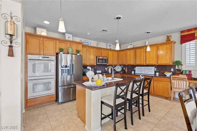 kitchen with hanging light fixtures, light tile patterned floors, stainless steel appliances, a kitchen island with sink, and decorative backsplash