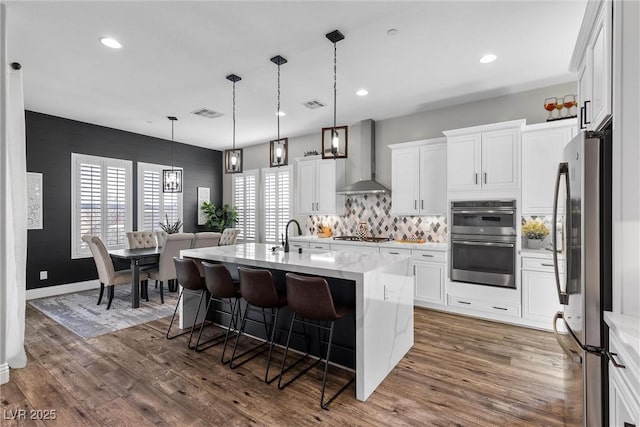 kitchen featuring visible vents, wood finished floors, stainless steel appliances, wall chimney range hood, and a kitchen bar