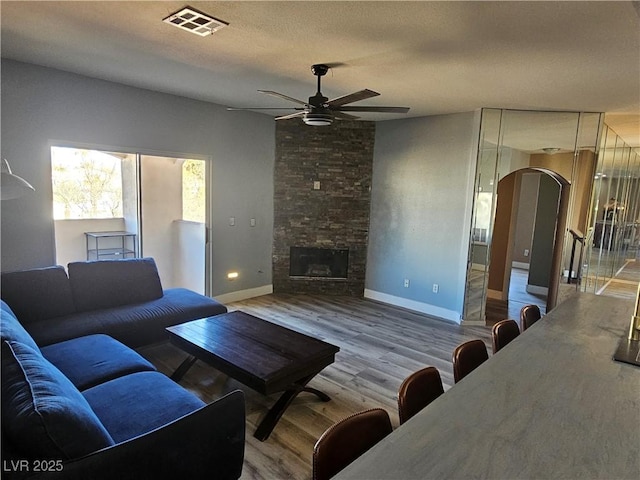 living room with ceiling fan, a stone fireplace, light hardwood / wood-style flooring, and a textured ceiling