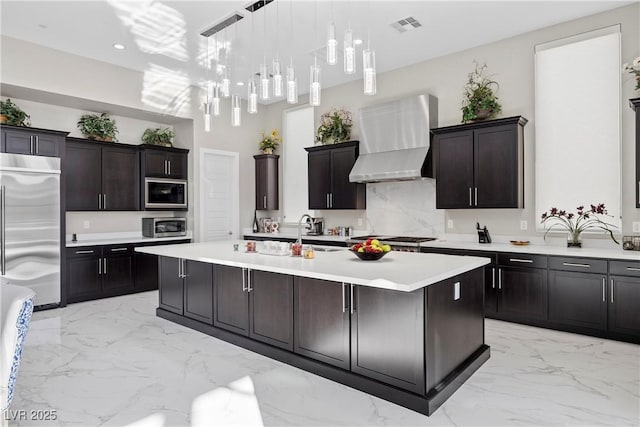 kitchen with built in appliances, an island with sink, dark brown cabinets, and wall chimney range hood