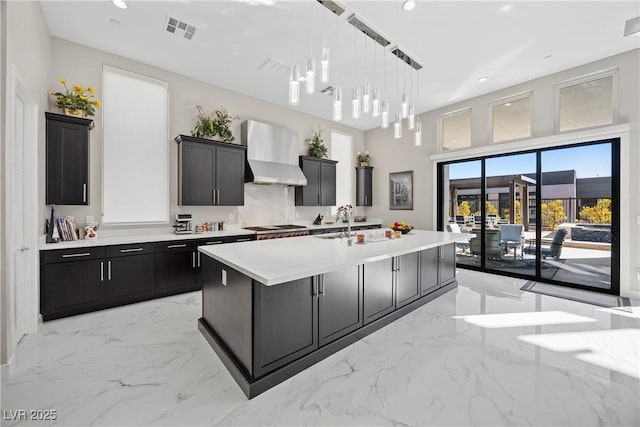 kitchen featuring wall chimney range hood, backsplash, an island with sink, decorative light fixtures, and stainless steel gas stovetop