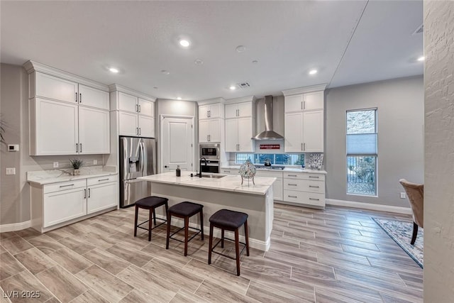 kitchen featuring appliances with stainless steel finishes, sink, white cabinets, a kitchen island with sink, and wall chimney range hood