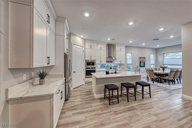 kitchen with wall chimney exhaust hood, sink, light stone counters, a center island with sink, and stainless steel appliances