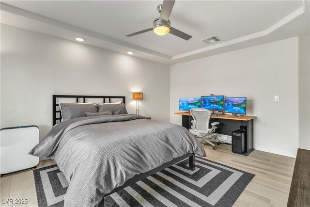 bedroom with ceiling fan, a tray ceiling, built in desk, and light wood-type flooring