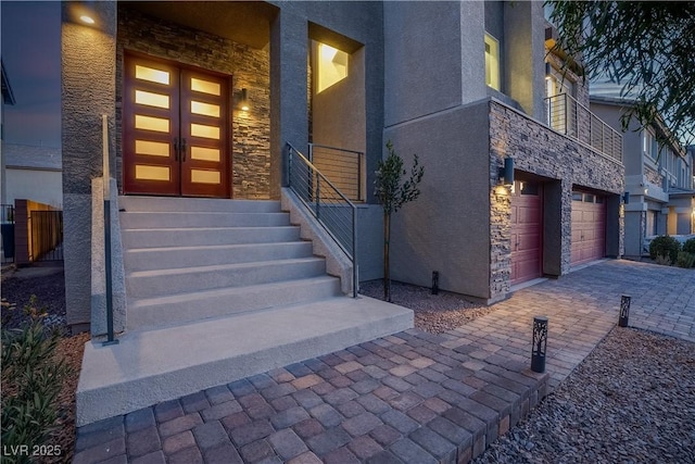 entrance to property with a garage, stone siding, decorative driveway, and stucco siding