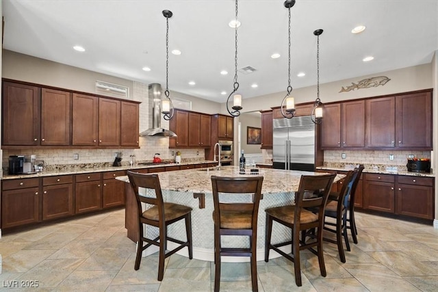 kitchen with a breakfast bar, hanging light fixtures, a kitchen island with sink, stainless steel appliances, and wall chimney range hood