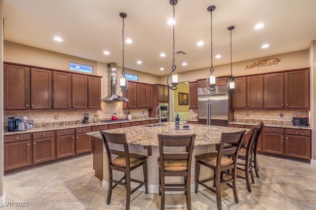 kitchen with wall chimney range hood, a breakfast bar area, appliances with stainless steel finishes, a center island with sink, and decorative light fixtures