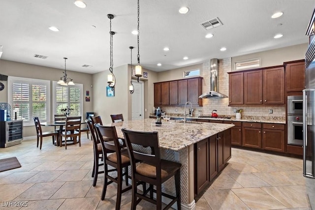 kitchen with decorative light fixtures, an island with sink, sink, a breakfast bar area, and wall chimney exhaust hood