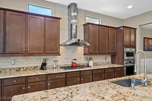 kitchen with wall chimney exhaust hood, sink, light stone counters, stainless steel appliances, and decorative backsplash