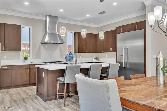 kitchen featuring wall chimney exhaust hood, stainless steel built in refrigerator, a kitchen bar, crown molding, and pendant lighting