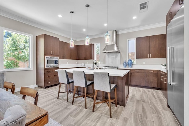 kitchen with decorative light fixtures, sink, built in appliances, dark brown cabinets, and wall chimney range hood