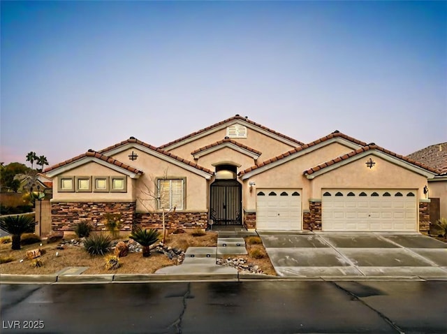 mediterranean / spanish-style house featuring a garage, driveway, stone siding, a tiled roof, and stucco siding