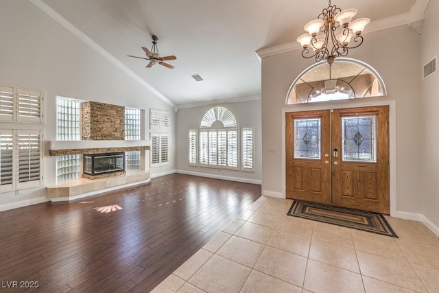 entryway with ceiling fan with notable chandelier, high vaulted ceiling, ornamental molding, a stone fireplace, and light wood-type flooring