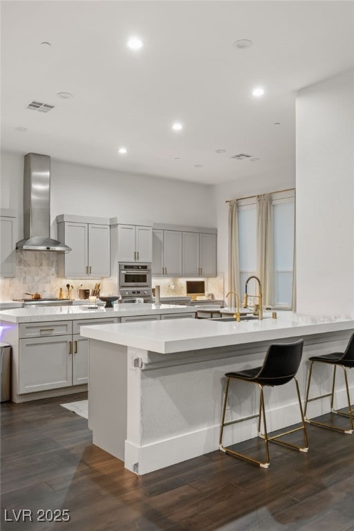 kitchen with an island with sink, a breakfast bar, dark wood-type flooring, and wall chimney exhaust hood