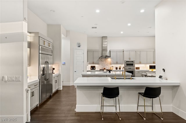 kitchen with a breakfast bar, sink, backsplash, stainless steel appliances, and wall chimney range hood