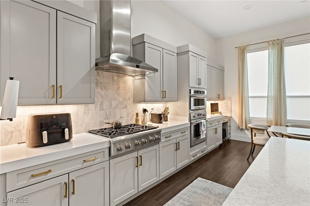 kitchen with wall chimney range hood, dark wood-type flooring, gray cabinets, stainless steel appliances, and tasteful backsplash