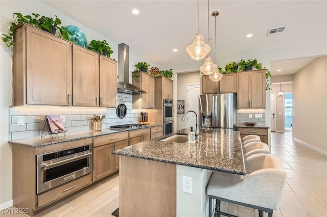 kitchen with sink, hanging light fixtures, stainless steel appliances, a kitchen island with sink, and wall chimney range hood