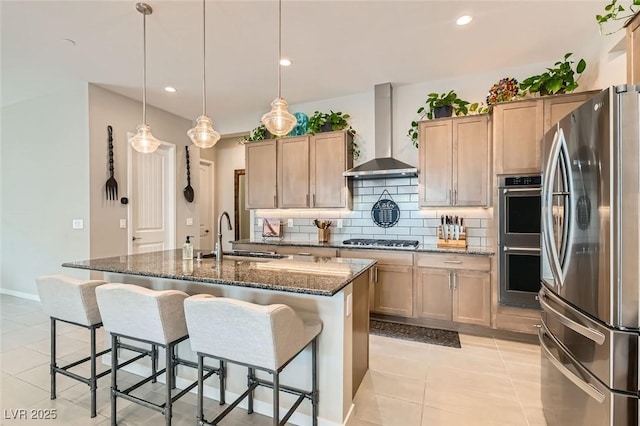 kitchen with appliances with stainless steel finishes, decorative light fixtures, an island with sink, dark stone counters, and wall chimney exhaust hood