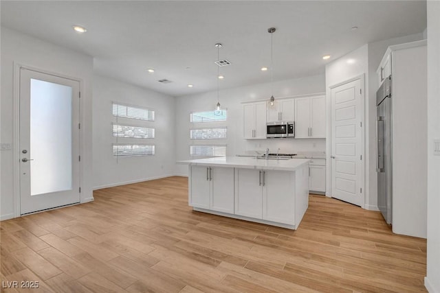 kitchen with hanging light fixtures, appliances with stainless steel finishes, a kitchen island with sink, light hardwood / wood-style floors, and white cabinets