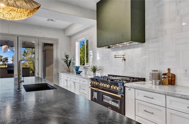 kitchen with visible vents, white cabinets, range with two ovens, wall chimney exhaust hood, and a sink