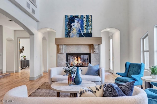 living room with arched walkways, light wood-style flooring, a high ceiling, a glass covered fireplace, and baseboards