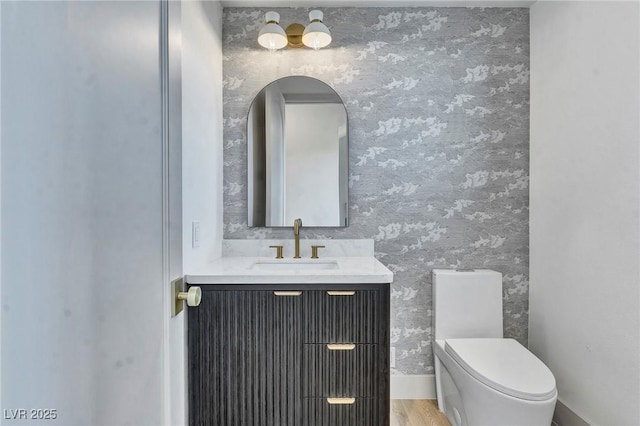 half bath with wood finished floors, vanity, toilet, and wallpapered walls