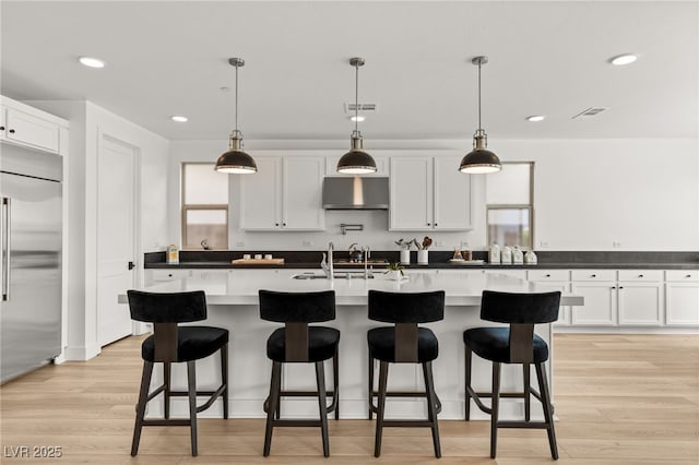 kitchen featuring built in refrigerator, hanging light fixtures, a center island with sink, and white cabinets