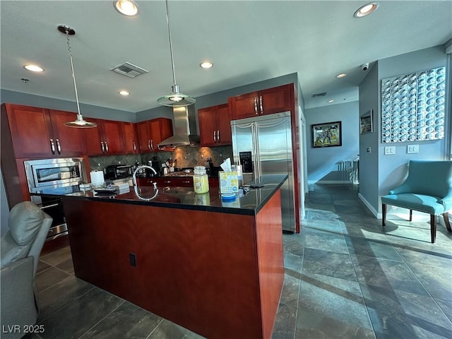 kitchen with built in appliances, hanging light fixtures, an island with sink, and wall chimney exhaust hood