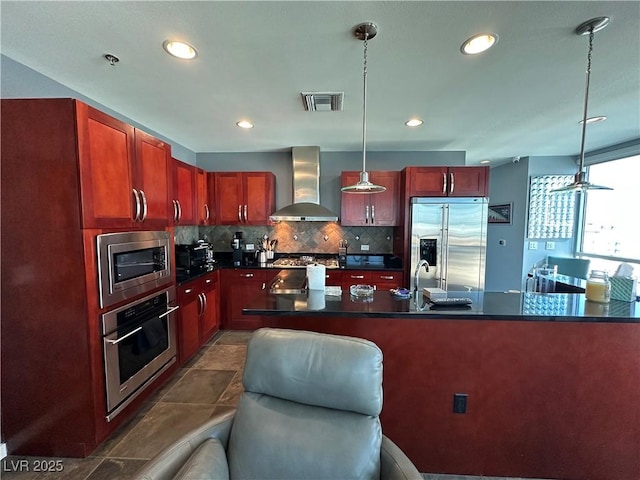 kitchen featuring pendant lighting, built in appliances, decorative backsplash, and wall chimney range hood