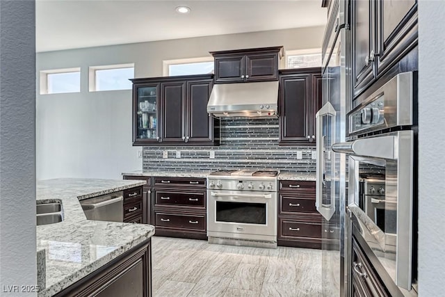 kitchen featuring under cabinet range hood, dark brown cabinets, appliances with stainless steel finishes, light stone countertops, and tasteful backsplash