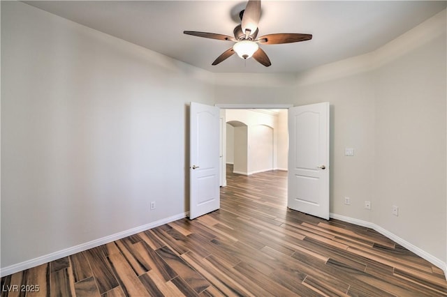 empty room featuring arched walkways, dark wood-style floors, a ceiling fan, and baseboards