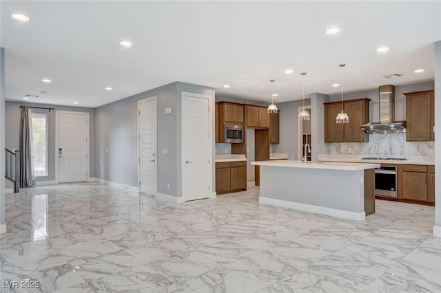 kitchen featuring a center island with sink, hanging light fixtures, appliances with stainless steel finishes, wall chimney range hood, and backsplash