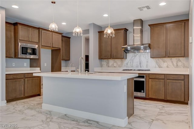 kitchen with appliances with stainless steel finishes, decorative light fixtures, tasteful backsplash, an island with sink, and wall chimney exhaust hood