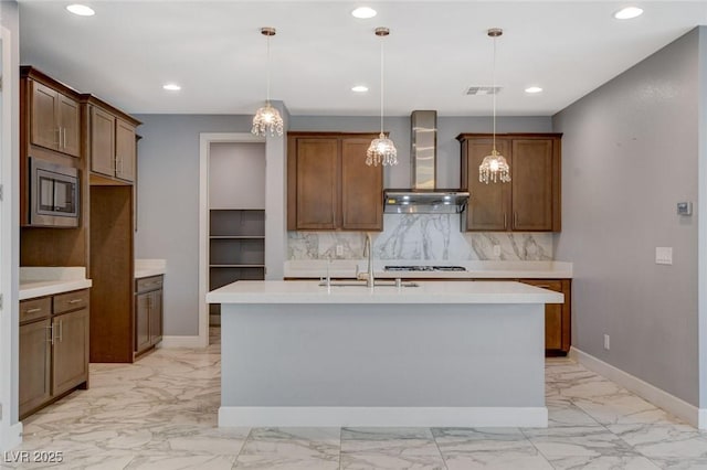 kitchen featuring stainless steel microwave, decorative backsplash, hanging light fixtures, a center island with sink, and wall chimney exhaust hood