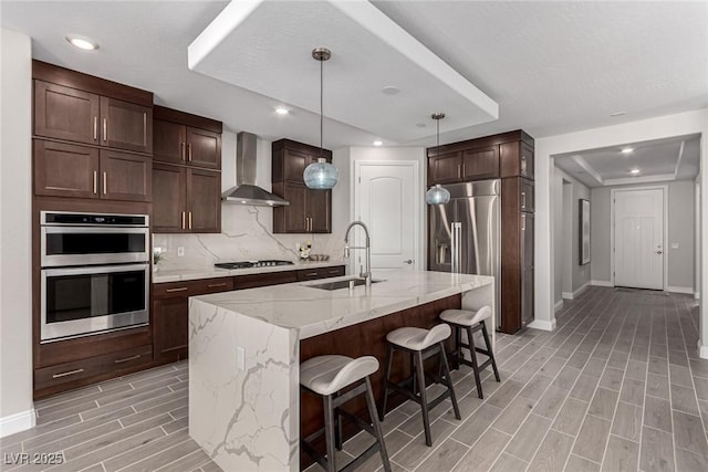 kitchen with wall chimney exhaust hood, sink, light stone counters, appliances with stainless steel finishes, and an island with sink