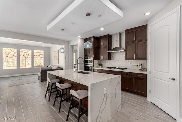 kitchen with sink, a kitchen island with sink, hanging light fixtures, light stone countertops, and wall chimney exhaust hood