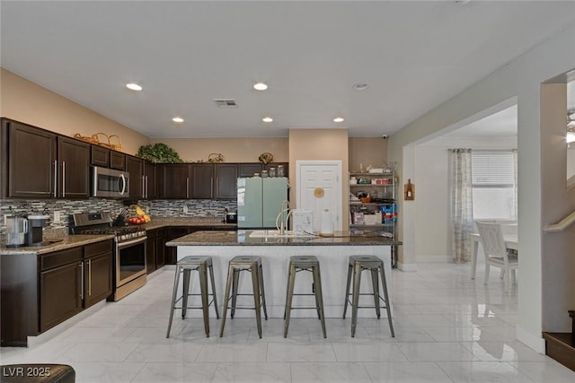 kitchen with a breakfast bar, appliances with stainless steel finishes, an island with sink, stone counters, and backsplash