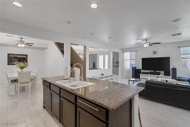 kitchen featuring dishwasher, sink, dark stone counters, dark brown cabinets, and a center island with sink