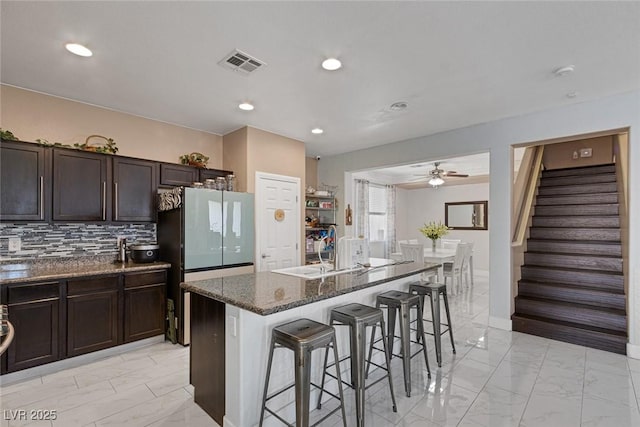 kitchen with sink, a breakfast bar area, dark stone countertops, fridge, and an island with sink