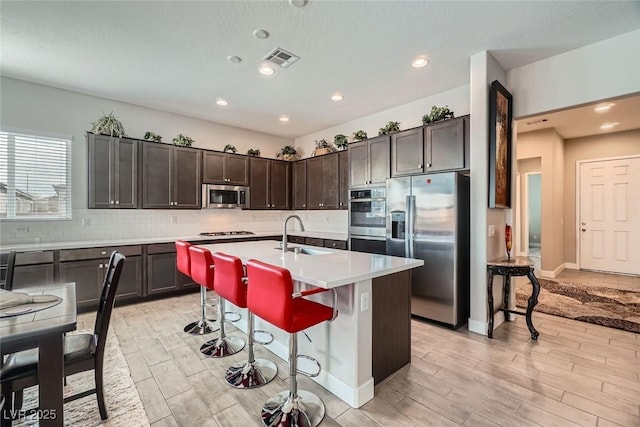 kitchen featuring a kitchen bar, decorative backsplash, a kitchen island with sink, dark brown cabinetry, and stainless steel appliances