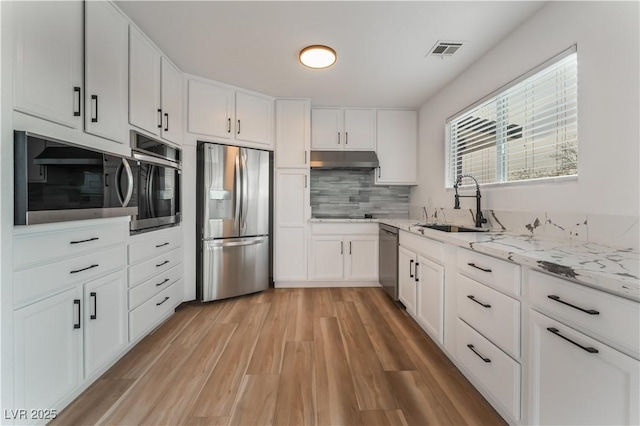 kitchen with appliances with stainless steel finishes, white cabinetry, sink, light stone countertops, and light wood-type flooring