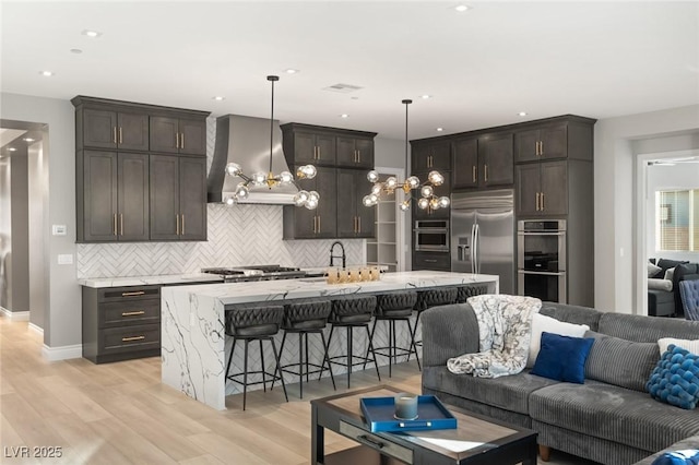 kitchen with a breakfast bar area, pendant lighting, stainless steel appliances, a kitchen island with sink, and wall chimney range hood