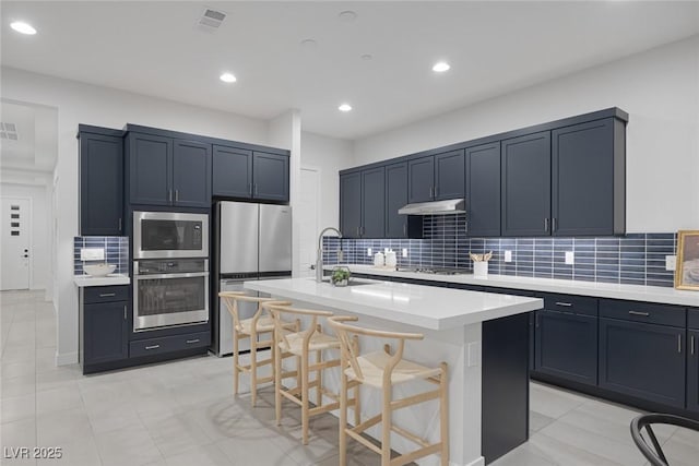 kitchen with sink, a center island with sink, stainless steel appliances, tasteful backsplash, and a kitchen bar