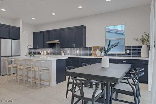 kitchen featuring stainless steel refrigerator, an island with sink, backsplash, a kitchen bar, and black electric stovetop