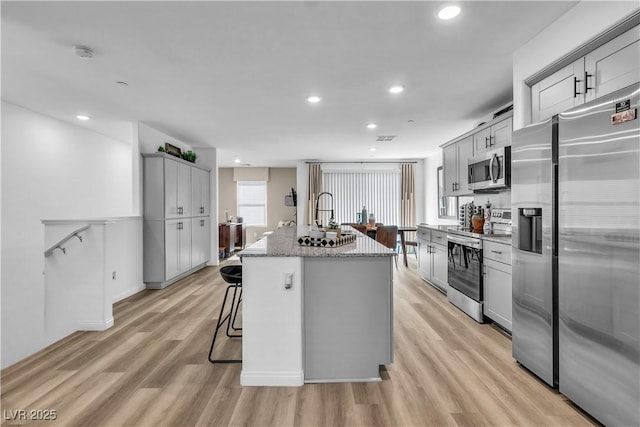 kitchen featuring appliances with stainless steel finishes, gray cabinetry, a kitchen island with sink, light stone counters, and light hardwood / wood-style flooring