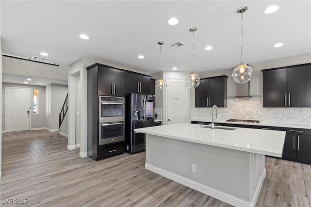 kitchen featuring sink, decorative light fixtures, a center island with sink, stainless steel appliances, and light hardwood / wood-style floors