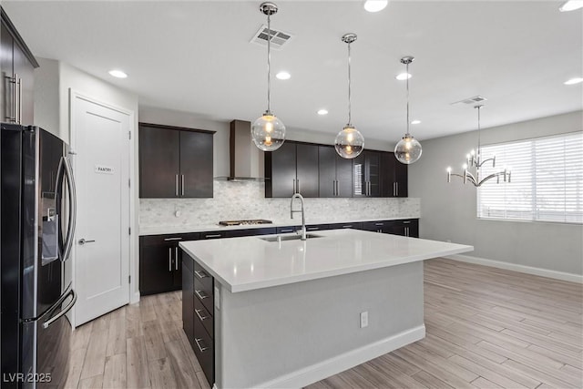 kitchen featuring decorative light fixtures, sink, a kitchen island with sink, black fridge with ice dispenser, and wall chimney exhaust hood