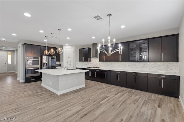 kitchen with sink, hanging light fixtures, a kitchen island with sink, stainless steel appliances, and wall chimney range hood