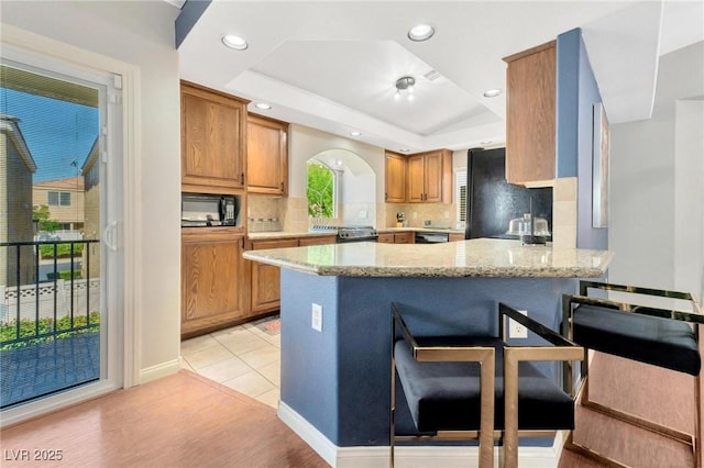 kitchen with backsplash, a tray ceiling, light stone countertops, black appliances, and kitchen peninsula