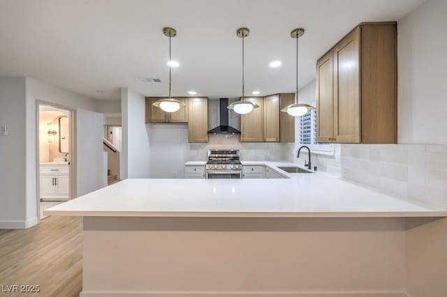 kitchen with stainless steel stove, a sink, light countertops, wall chimney range hood, and hanging light fixtures